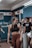 A young woman with braided hair is in a gym setting, wearing a black sports bra and brown leggings. She appears to be conversing with or listening to people around her. The background shows people engaged in a fitness environment, with gym equipment and motivational chalk writings on the wall.