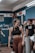 A young woman with braided hair is in a gym setting, wearing a black sports bra and brown leggings. She appears to be conversing with or listening to people around her. The background shows people engaged in a fitness environment, with gym equipment and motivational chalk writings on the wall.