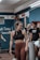 A young woman with braided hair is in a gym setting, wearing a black sports bra and brown leggings. She appears to be conversing with or listening to people around her. The background shows people engaged in a fitness environment, with gym equipment and motivational chalk writings on the wall.