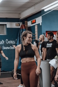 A young woman with braided hair is in a gym setting, wearing a black sports bra and brown leggings. She appears to be conversing with or listening to people around her. The background shows people engaged in a fitness environment, with gym equipment and motivational chalk writings on the wall.