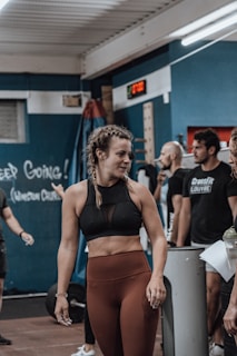 A young woman with braided hair is in a gym setting, wearing a black sports bra and brown leggings. She appears to be conversing with or listening to people around her. The background shows people engaged in a fitness environment, with gym equipment and motivational chalk writings on the wall.