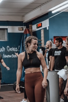 A young woman with braided hair is in a gym setting, wearing a black sports bra and brown leggings. She appears to be conversing with or listening to people around her. The background shows people engaged in a fitness environment, with gym equipment and motivational chalk writings on the wall.