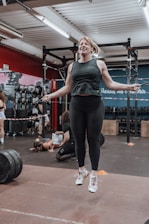 A woman is using a jump rope in a gym, surrounded by equipment such as weights and exercise mats. Several people are in the background, some engaged in exercise activities. The gym environment features red and blue walls and motivational quotes.