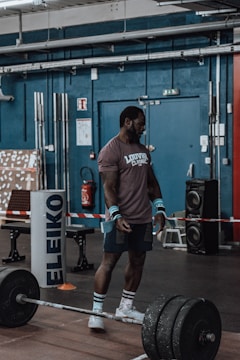 A man stands in a gym wearing athletic apparel, appearing to prepare for weightlifting. He is positioned near a barbell with multiple weight plates. The gym environment includes various fitness equipment, weights, and industrial-style decor.