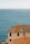 A picturesque seaside view featuring a large terracotta-roofed building with multiple windows. The building is situated on a coastal cliff, overlooking an expansive view of calm blue ocean water stretching to the horizon under a clear sky.