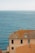 A picturesque seaside view featuring a large terracotta-roofed building with multiple windows. The building is situated on a coastal cliff, overlooking an expansive view of calm blue ocean water stretching to the horizon under a clear sky.