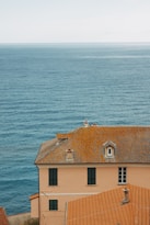 A picturesque seaside view featuring a large terracotta-roofed building with multiple windows. The building is situated on a coastal cliff, overlooking an expansive view of calm blue ocean water stretching to the horizon under a clear sky.