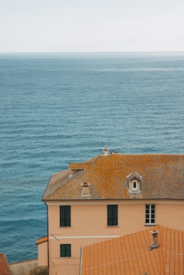 A picturesque seaside view featuring a large terracotta-roofed building with multiple windows. The building is situated on a coastal cliff, overlooking an expansive view of calm blue ocean water stretching to the horizon under a clear sky.
