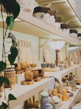 A neatly organized shelf displays a variety of wooden and woven kitchen and home decor items. The arrangement includes baskets, bowls, utensils, and decorative bags, surrounded by natural elements like leafy plants. The ambiance gives off a rustic and earthy feel.
