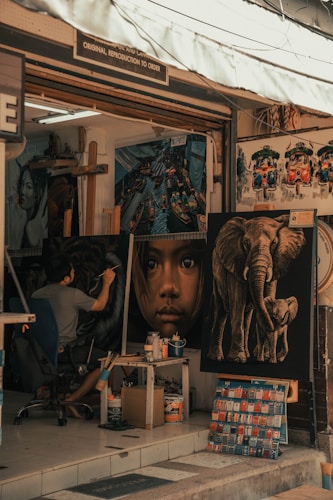 An art studio facade displaying several large paintings including a close-up portrait of a child, an elephant with its trunk curled, and colorful vehicles resembling tuk-tuks. Inside, an artist is seated, intently working on a painting, surrounded by art supplies and tools.