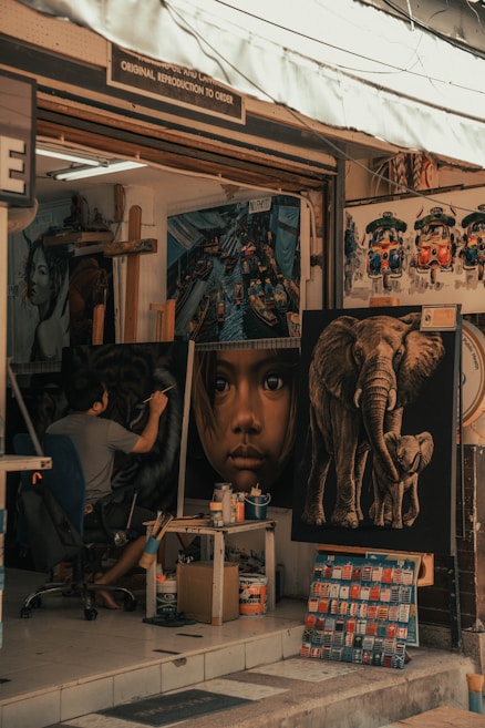 An art studio facade displaying several large paintings including a close-up portrait of a child, an elephant with its trunk curled, and colorful vehicles resembling tuk-tuks. Inside, an artist is seated, intently working on a painting, surrounded by art supplies and tools.