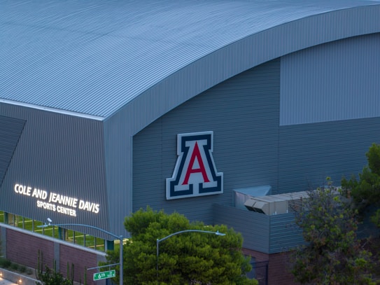 A large building with a corrugated metal exterior features a prominent logo displaying the letter 'A' in bold colors. Below the logo, the words 'Cole and Jeannie Davis Sports Center' are illuminated. Trees and a street sign are visible in the foreground.