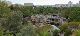 A scenic view of a zoo or wildlife sanctuary featuring a large enclosure with wooden structures, possibly for primates. There are lush green trees surrounding the enclosure and a clear pathway is visible. Beyond the trees, there are multiple high-rise buildings indicating an urban setting.