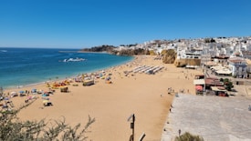 A sunny beach scene with clear blue skies and a vibrant blue ocean. The sandy beach is populated with colorful umbrellas and people enjoying the sun and sea. A row of white buildings is visible on the right, nestled against the cliffs, suggesting a coastal town. The scene evokes a lively, summery atmosphere.