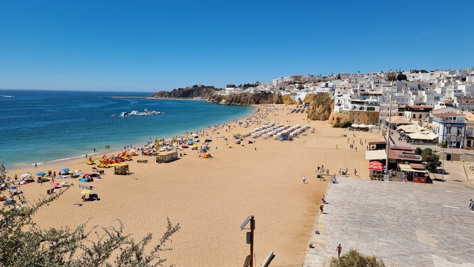 A sunny beach scene with clear blue skies and a vibrant blue ocean. The sandy beach is populated with colorful umbrellas and people enjoying the sun and sea. A row of white buildings is visible on the right, nestled against the cliffs, suggesting a coastal town. The scene evokes a lively, summery atmosphere.