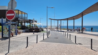A seaside promenade with a pathway leading towards a viewpoint overlooking the ocean. On the left, there's a small restaurant with outdoor seating, offering gelato and pizza. Several people are sitting on benches or walking near the ocean view. A large, curved wooden canopy provides shade along the walkway.