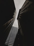 Tall buildings are captured from a low-angle perspective, creating a dramatic and imposing view as they reach up towards a cloudy sky. The structures appear dark in contrast to the pale backdrop, accentuating their geometric lines.