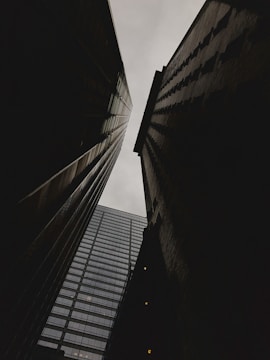 Tall buildings are captured from a low-angle perspective, creating a dramatic and imposing view as they reach up towards a cloudy sky. The structures appear dark in contrast to the pale backdrop, accentuating their geometric lines.