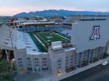 An aerial view of a large football stadium with a lush green field, surrounded by empty stands and a prominent logo on a large structure. The stadium is located in an urban area with mountain ranges visible in the background under a partly cloudy sky.