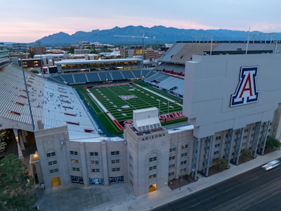 An aerial view of a large football stadium with a lush green field, surrounded by empty stands and a prominent logo on a large structure. The stadium is located in an urban area with mountain ranges visible in the background under a partly cloudy sky.