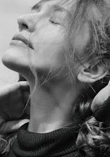 A black and white portrait of a person with eyes closed, head tilted upward, and hair flowing around the face. The expression conveys tranquility, and the person is gently touching their neck with both hands.