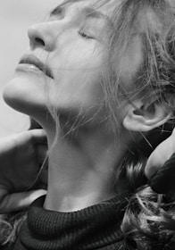 A black and white portrait of a person with eyes closed, head tilted upward, and hair flowing around the face. The expression conveys tranquility, and the person is gently touching their neck with both hands.