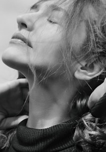 A black and white portrait of a person with eyes closed, head tilted upward, and hair flowing around the face. The expression conveys tranquility, and the person is gently touching their neck with both hands.