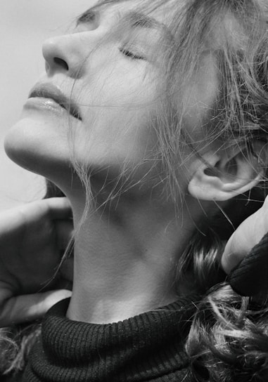 A black and white portrait of a person with eyes closed, head tilted upward, and hair flowing around the face. The expression conveys tranquility, and the person is gently touching their neck with both hands.