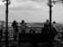 A group of people stand on a platform overlooking a panoramic view. The scene includes several individuals, with their backs turned, looking out over a cityscape under a cloudy sky. The architecture of the platform features decorative wrought-iron detailing.