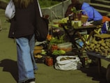 A street market scene features a variety of vegetables and goods displayed on tables and in crates. Fresh produce like potatoes, tomatoes, and greens are visible, alongside jars of preserved items. People are casually browsing and selling items, adding a lively element to the setting.