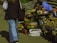 A street market scene features a variety of vegetables and goods displayed on tables and in crates. Fresh produce like potatoes, tomatoes, and greens are visible, alongside jars of preserved items. People are casually browsing and selling items, adding a lively element to the setting.