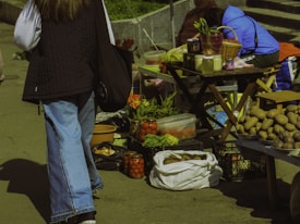 A street market scene features a variety of vegetables and goods displayed on tables and in crates. Fresh produce like potatoes, tomatoes, and greens are visible, alongside jars of preserved items. People are casually browsing and selling items, adding a lively element to the setting.