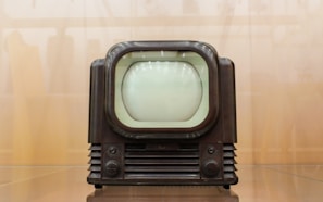 A vintage television set with a rounded screen and a dark brown, wooden exterior. The front features two control knobs and grill-like designs below the screen. The background is a blurred, light brown hue, giving the image a nostalgic feel.