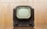 A vintage television set with a rounded screen and a dark brown, wooden exterior. The front features two control knobs and grill-like designs below the screen. The background is a blurred, light brown hue, giving the image a nostalgic feel.