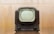 A vintage television set with a rounded screen and a dark brown, wooden exterior. The front features two control knobs and grill-like designs below the screen. The background is a blurred, light brown hue, giving the image a nostalgic feel.
