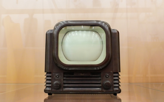 A vintage television set with a rounded screen and a dark brown, wooden exterior. The front features two control knobs and grill-like designs below the screen. The background is a blurred, light brown hue, giving the image a nostalgic feel.