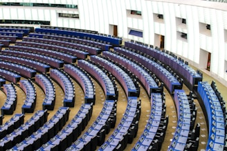 A large, modern auditorium with curved rows of desks and seats, arranged in a semicircular pattern. The seating is predominantly blue and black, with each desk having its own microphone and electronic device. The walls feature vertical panels that appear to be illuminated or reflective, contributing to a clean, contemporary look.