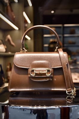 A luxurious brown leather handbag with a polished gold clasp and buckle design is displayed prominently. The structured bag features a top handle and a detachable strap. The background shows a boutique setting with shelves of other handbags and luxury items, softly illuminated with spotlights.