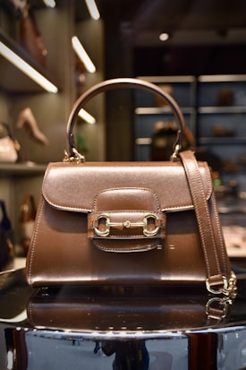 A luxurious brown leather handbag with a polished gold clasp and buckle design is displayed prominently. The structured bag features a top handle and a detachable strap. The background shows a boutique setting with shelves of other handbags and luxury items, softly illuminated with spotlights.