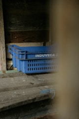 A set of blue plastic crates with the label 'INDUVECA 2' is placed in a dimly lit, rustic environment. The wooden walls and surfaces have a weathered appearance, adding to the rustic feel.