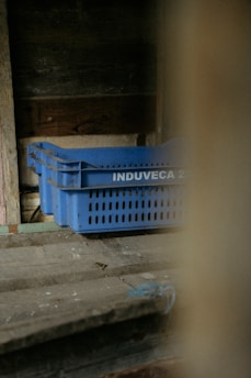 A set of blue plastic crates with the label 'INDUVECA 2' is placed in a dimly lit, rustic environment. The wooden walls and surfaces have a weathered appearance, adding to the rustic feel.
