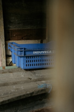 A set of blue plastic crates with the label 'INDUVECA 2' is placed in a dimly lit, rustic environment. The wooden walls and surfaces have a weathered appearance, adding to the rustic feel.