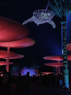 A nighttime scene features large, circular structures illuminated with red lighting. A crowd of people stands beneath, with palm trees adding to the ambiance. Overhead, a large, illuminated sea turtle sculpture composed of small lights creates a mystical atmosphere.