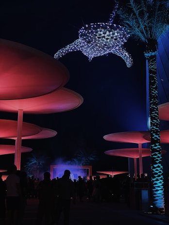 A nighttime scene features large, circular structures illuminated with red lighting. A crowd of people stands beneath, with palm trees adding to the ambiance. Overhead, a large, illuminated sea turtle sculpture composed of small lights creates a mystical atmosphere.