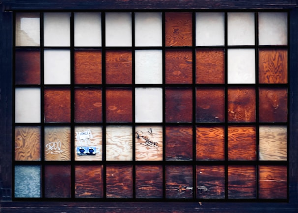 A window grid with several rectangular glass panels arranged in a geometric pattern. Some of the panels are frosted or opaque, while others have a warm, wooden texture. A few panels display graffiti and stickers, adding an element of urban art.