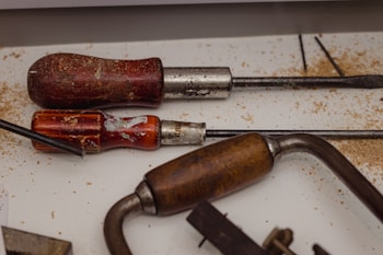 A collection of woodworking tools with wooden and metal handles lies on a surface sprinkled with wood shavings. The tools include chisels and a coping saw, displaying signs of wear and use.