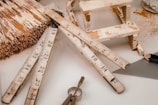 Assorted construction and woodworking tools arranged on a surface, including a folding ruler, a paintbrush with visible paint splatters, a small wooden stool or framework, a triangular ruler, a pencil, and a key. The objects appear well-used, suggesting frequent usage in woodworking or DIY projects.