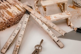 Assorted construction and woodworking tools arranged on a surface, including a folding ruler, a paintbrush with visible paint splatters, a small wooden stool or framework, a triangular ruler, a pencil, and a key. The objects appear well-used, suggesting frequent usage in woodworking or DIY projects.
