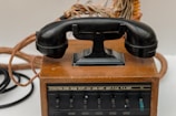 An antique rotary-style telephone rests on a wooden base with visible cords and a series of labeled switches, possibly indicating a multi-line or switchboard functionality.