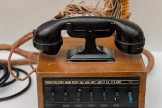 An antique rotary-style telephone rests on a wooden base with visible cords and a series of labeled switches, possibly indicating a multi-line or switchboard functionality.